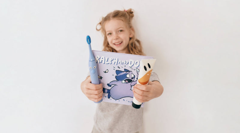 Mädchen zeigt Schallzahnbürste, Kinderzahnpasta und Zahnputzbuch